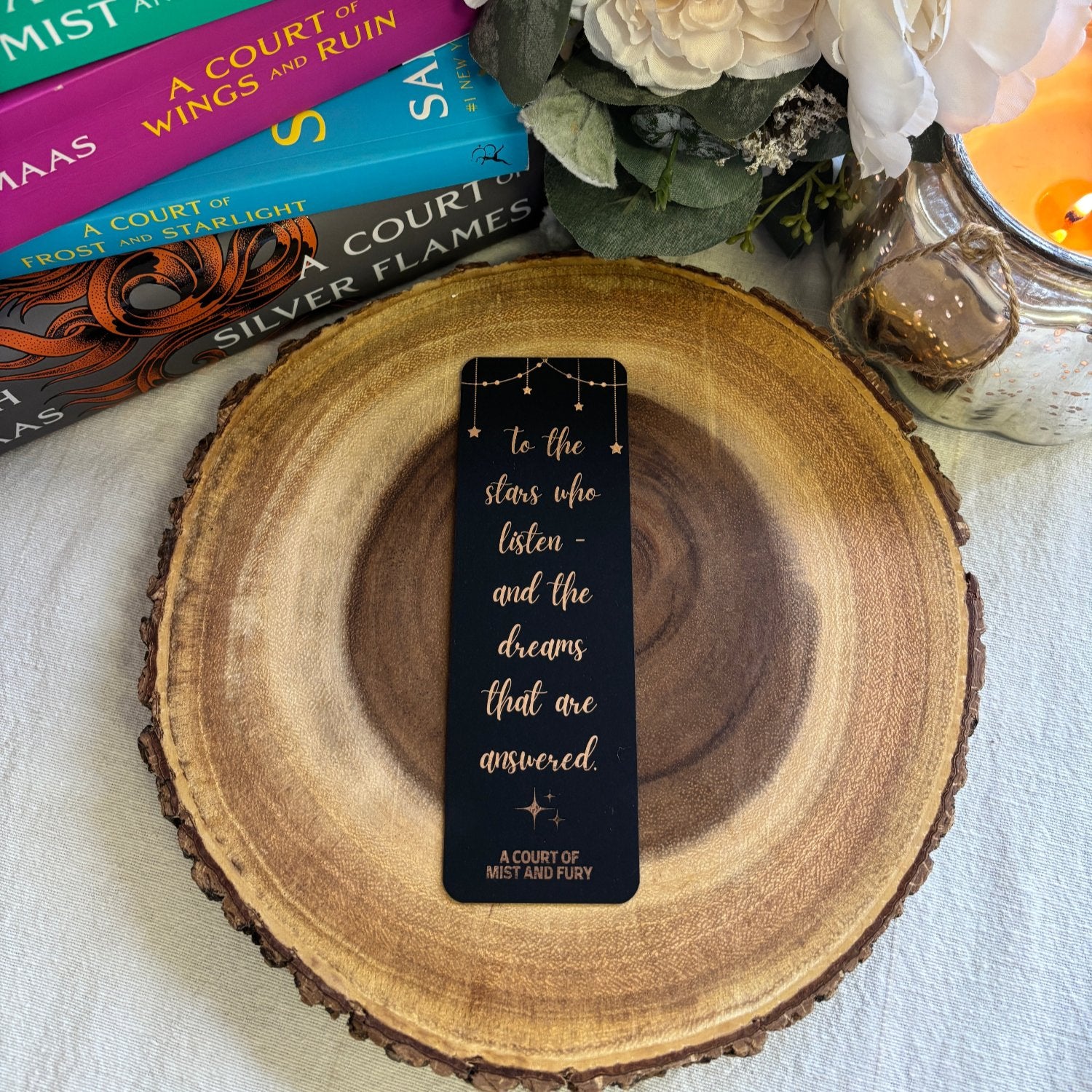 rose gold foil bookmark is displayed on a round piece of wood with a stack of books, flowers and candle in the background