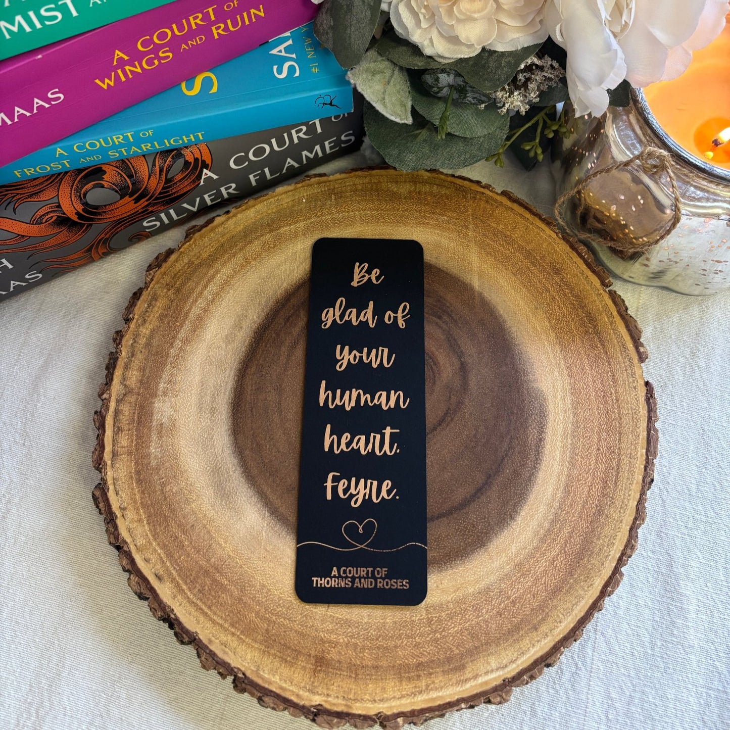 black bookmark with rose gold writing that reads "be glad of your human heart Feyre". bookmark sits on a wooden block, with a stack of books, flowers and candle in the background